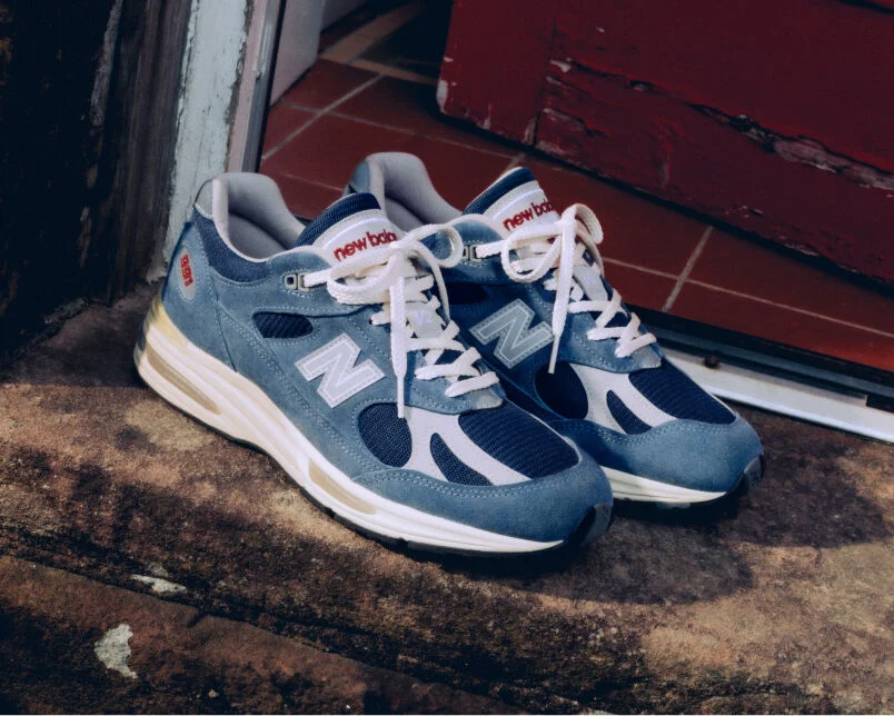 A pair of New Balance Made in UK sneakers sitting on an old stone doorstep next to a red door. 