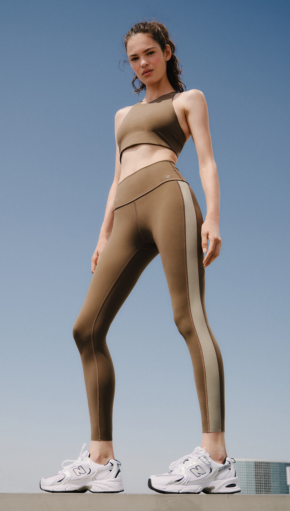 An individual wearing a brown sports bra and brown leggings standing on a concrete ledge in front of blue sky. 
