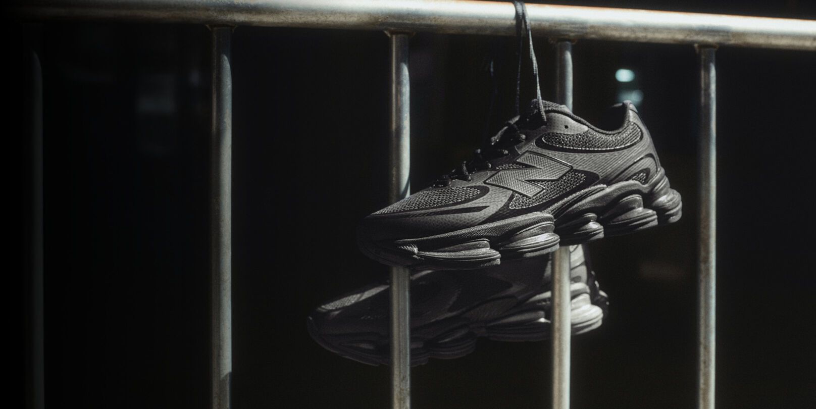 Black New Balance sneakers hanging by laces over a metal railing in dramatic lighting.