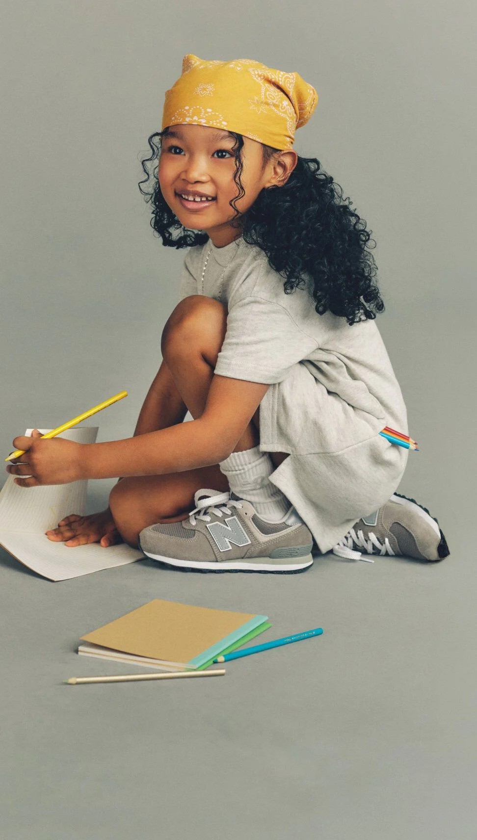 Child crouching on the floor writing in a notebook with colored pencils, wearing a yellow bandana, gray outfit, white socks, and gray New Balance sneakers against a plain background.