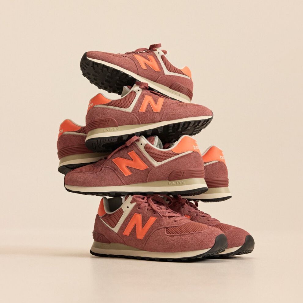 Stack of brick‑red New Balance sneakers with orange accents arranged in a balanced tower against a beige background.