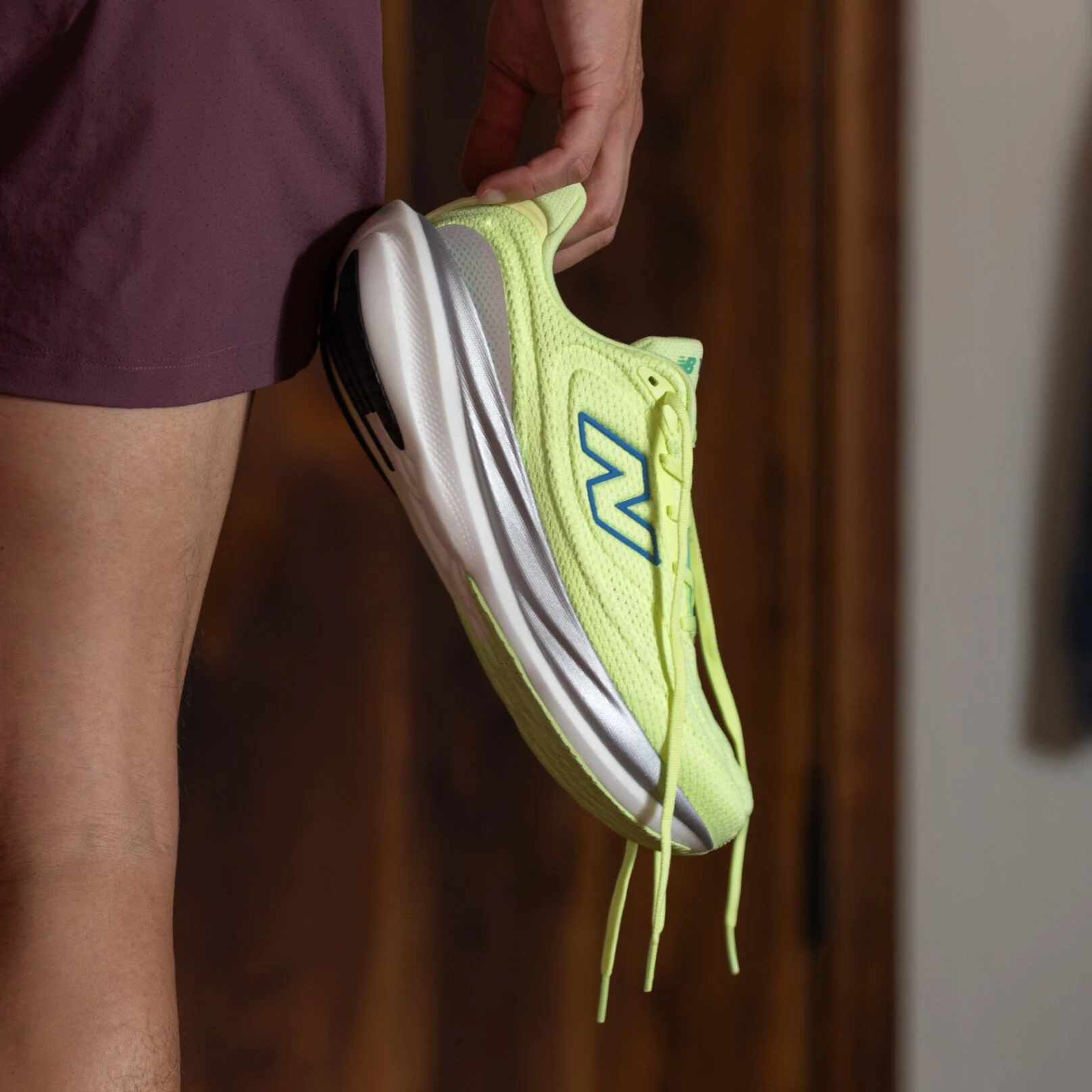 Close-up of a bright yellow-green New Balance running shoe with blue logo and white sole, held by a person wearing maroon shorts. 