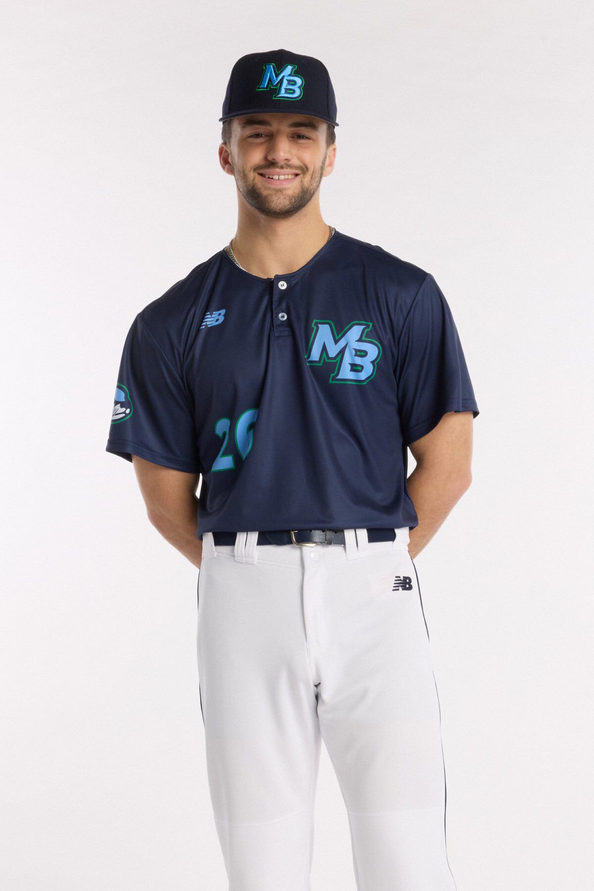 Athlete wearing a navy baseball jersey with teal &ldquo;MB&rdquo; logo, matching navy cap, and white baseball pants, standing against a light background.