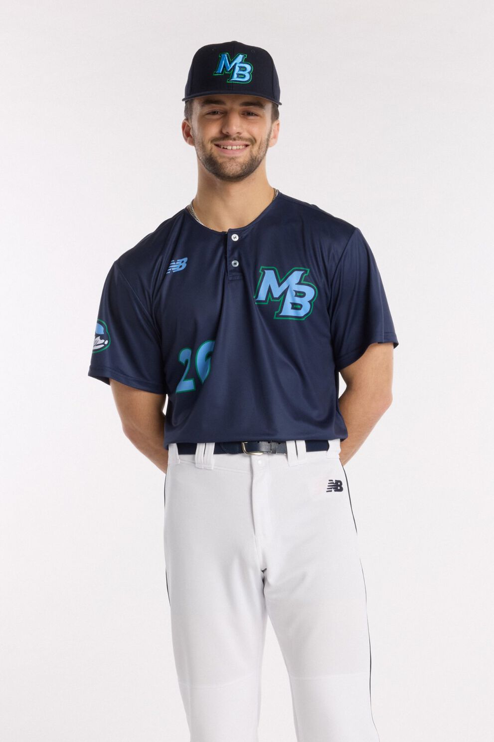 Athlete wearing a navy baseball jersey with teal &ldquo;MB&rdquo; logo, matching navy cap, and white baseball pants, standing against a light background.