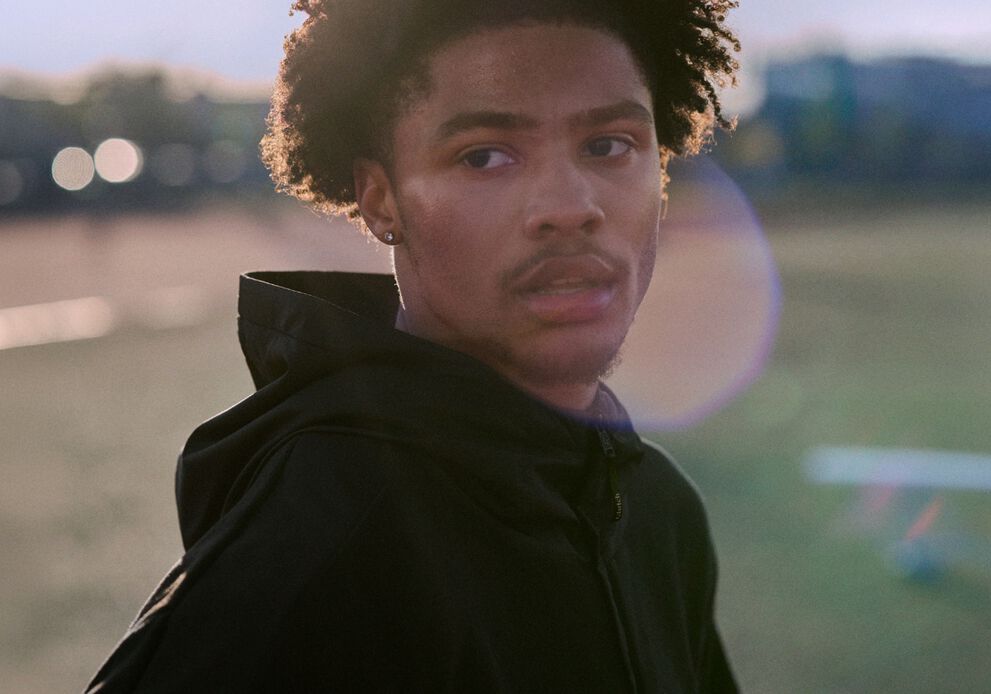 Athlete wearing a black hoodie standing on an outdoor track and field at dusk.