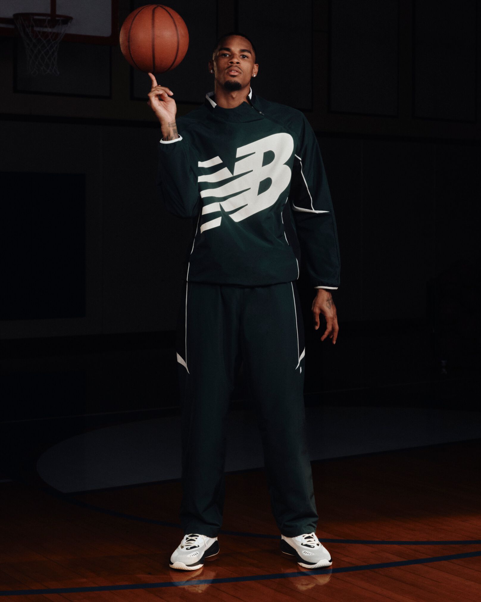 Person standing on a basketball court in a black New Balance tracksuit, balancing a basketball on one hand while wearing white athletic sneakers.