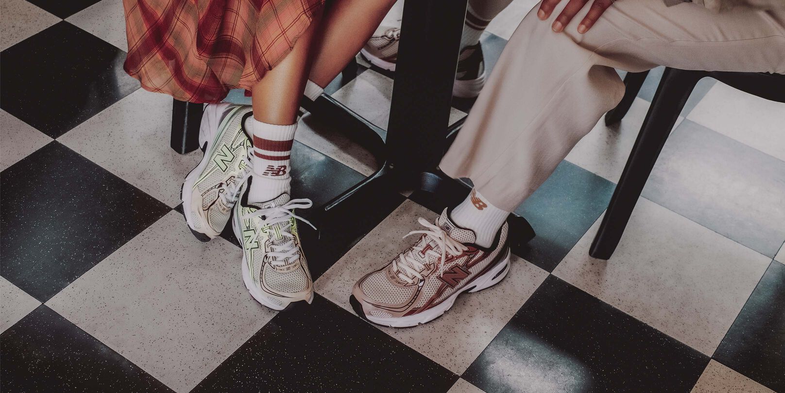Knee-down image of a male and female model sitting at a table wearing the New Balance 740 sneakers in the brown metallic colorway and the mint flash green colorway.