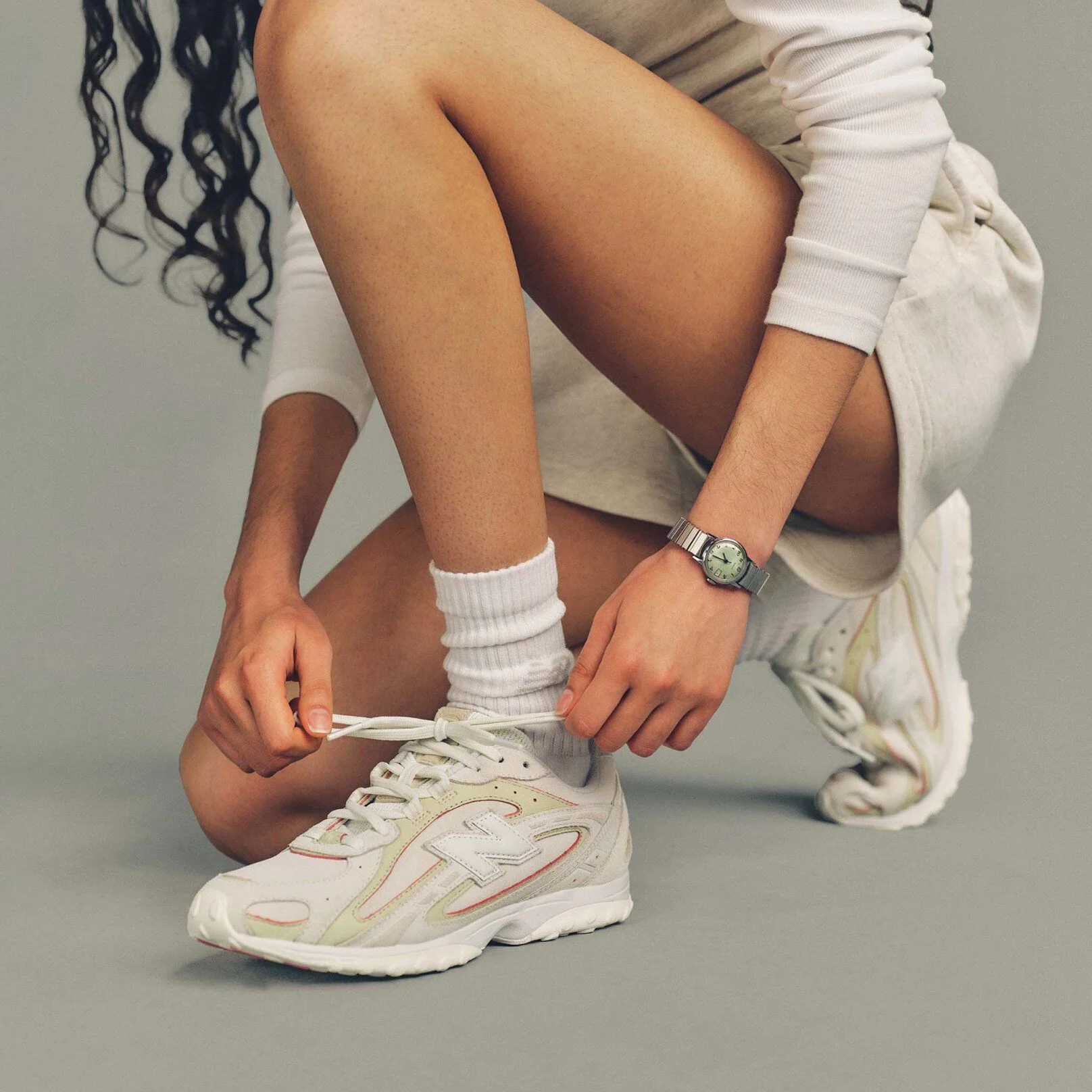 A person kneels on the ground while tying the laces of a white New Balance sneaker with pastel accents.