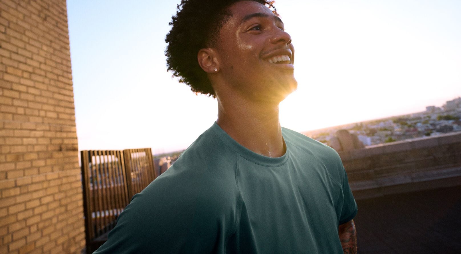 Athlete in a teal performance T-shirt standing on a rooftop training area at sunset.