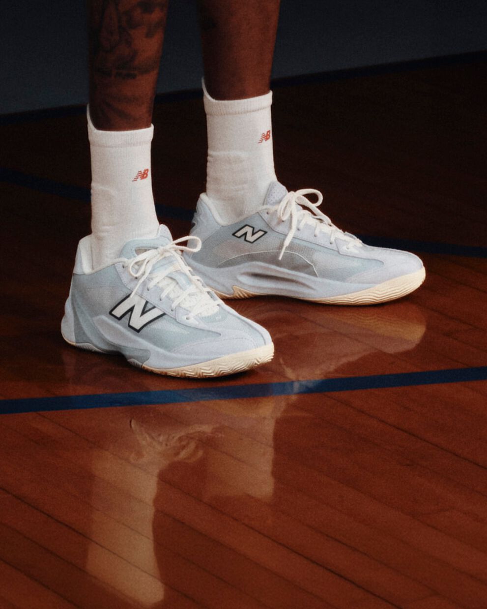 Light blue and white New Balance basketball shoes worn with white crew socks on a polished indoor basketball court.