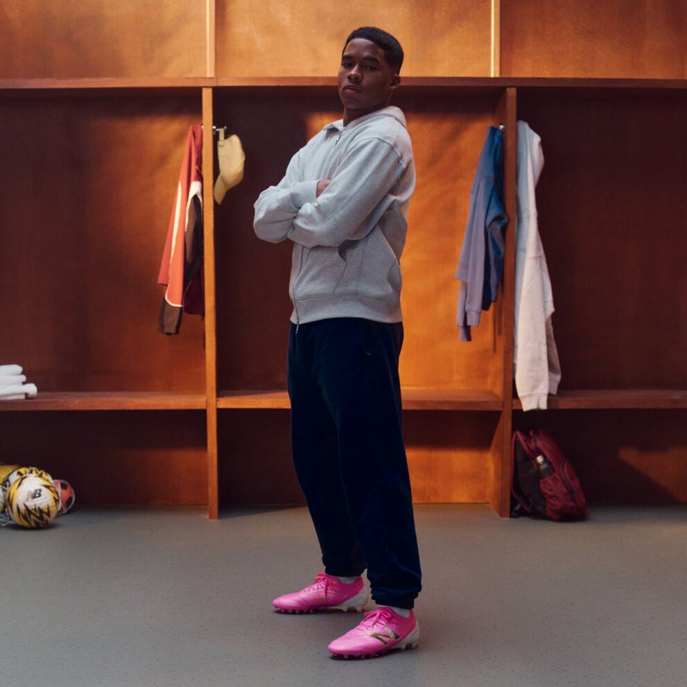 Endrick de pie en un vestuario de madera con los brazos cruzados, con una sudadera gris claro con cremallera, pantalones deportivos oscuros y botas de f&uacute;tbol rosa brillante; taquillas abiertas detr&aacute;s exponen ropa colgada, ropa doblada, una mochila y balones de f&uacute;tbol en el suelo.