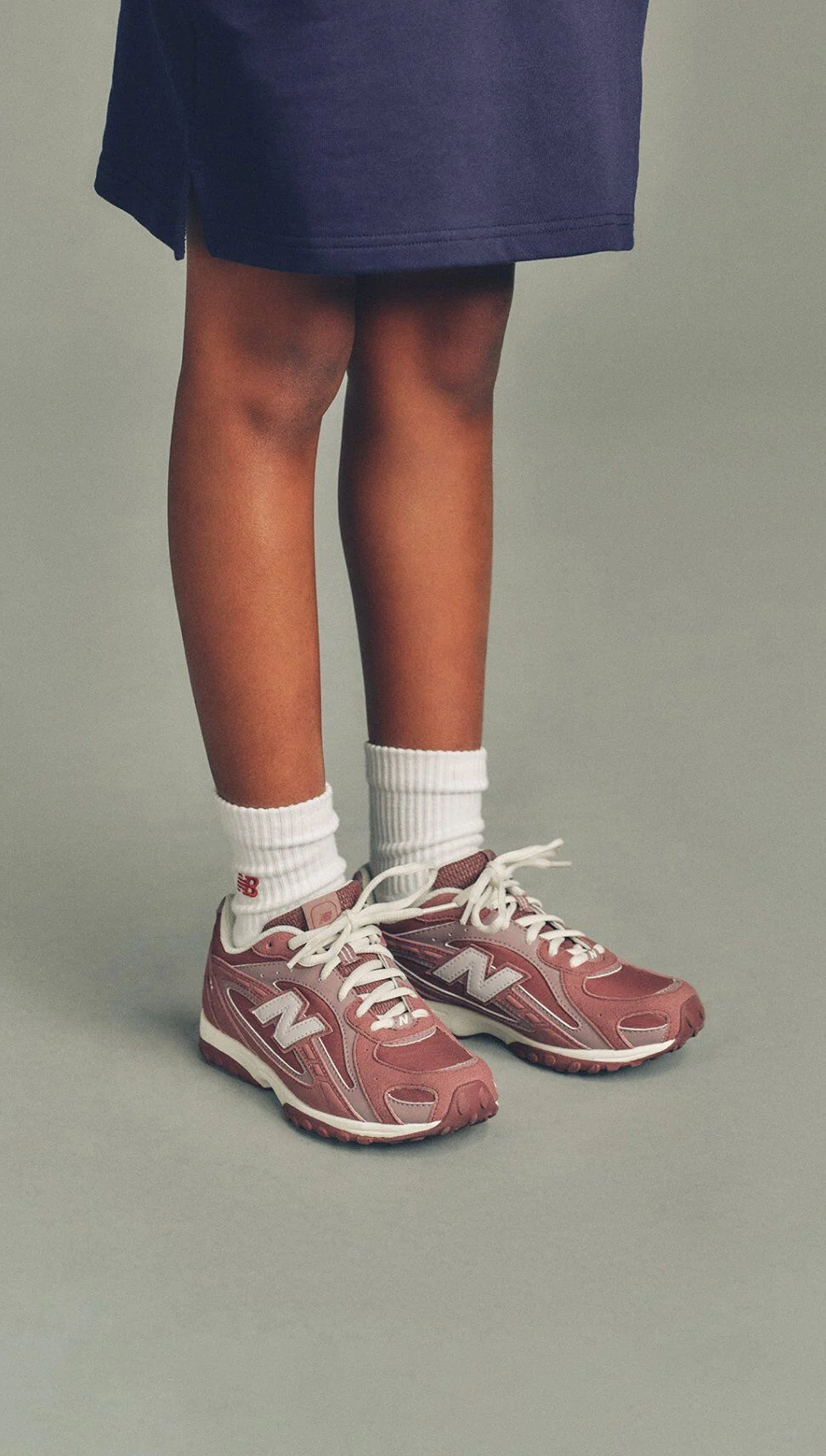 Close-up of a child wearing white socks and pink New Balance sneakers standing against a neutral background.