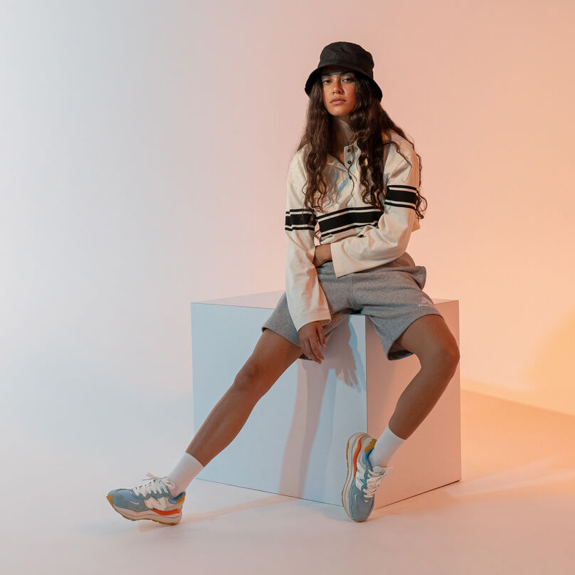 Woman sitting on box in multi-colored studio environment wearing a hat, white sweatshirt, grey shorts, and 57/40 sneakers.
