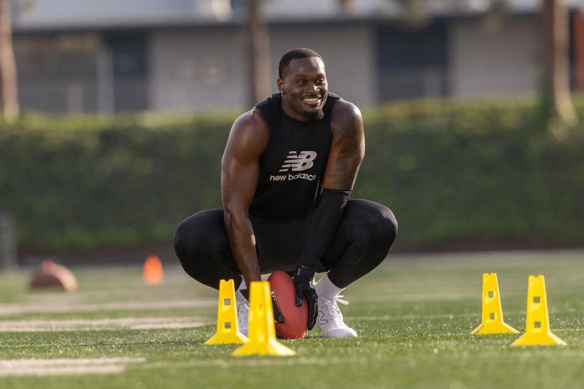 Young man crouches on a grass field holding a football between yellow cones during an agility setup, wearing black training apparel and white cleats.