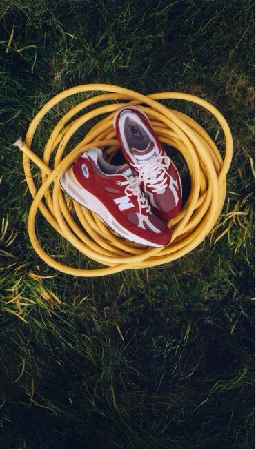 A pair of New Balance Made in UK sneakers sitting on a yellow coiled hose on top of a lush grass lawn.