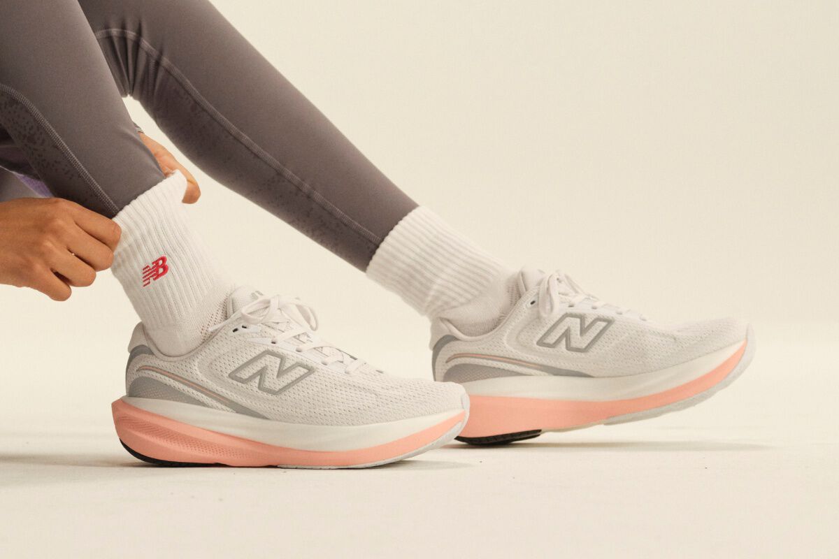 Close-up of white New Balance running shoes with pink midsoles paired with white crew socks and gray leggings in a clean studio setting.