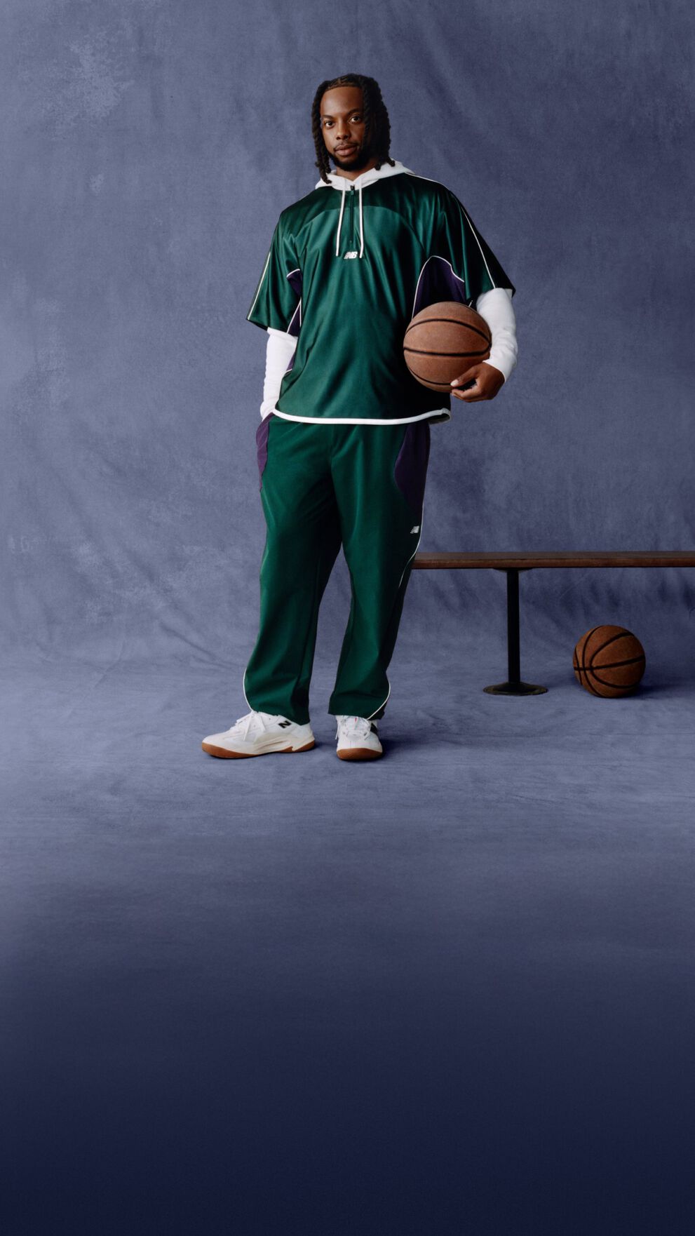 An individual stands in a studio setting wearing a dark green athletic outfit with white and purple accents, including a short‑sleeve top layered over a long‑sleeve shirt and matching track pants.