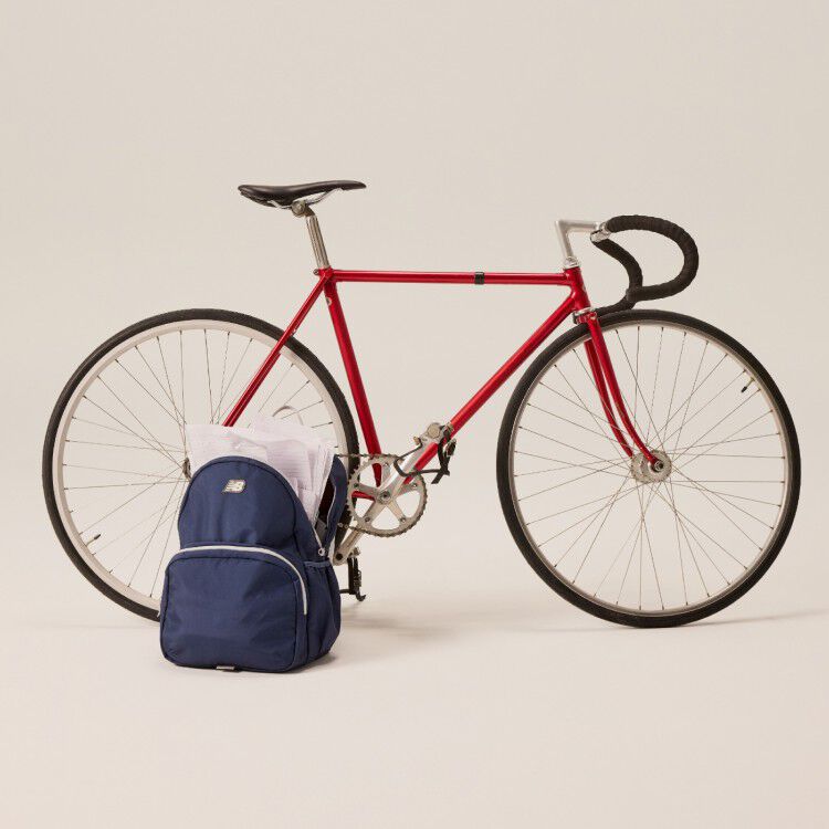 A red bicycle next to a blue New Balance backpack in front of a beige background.