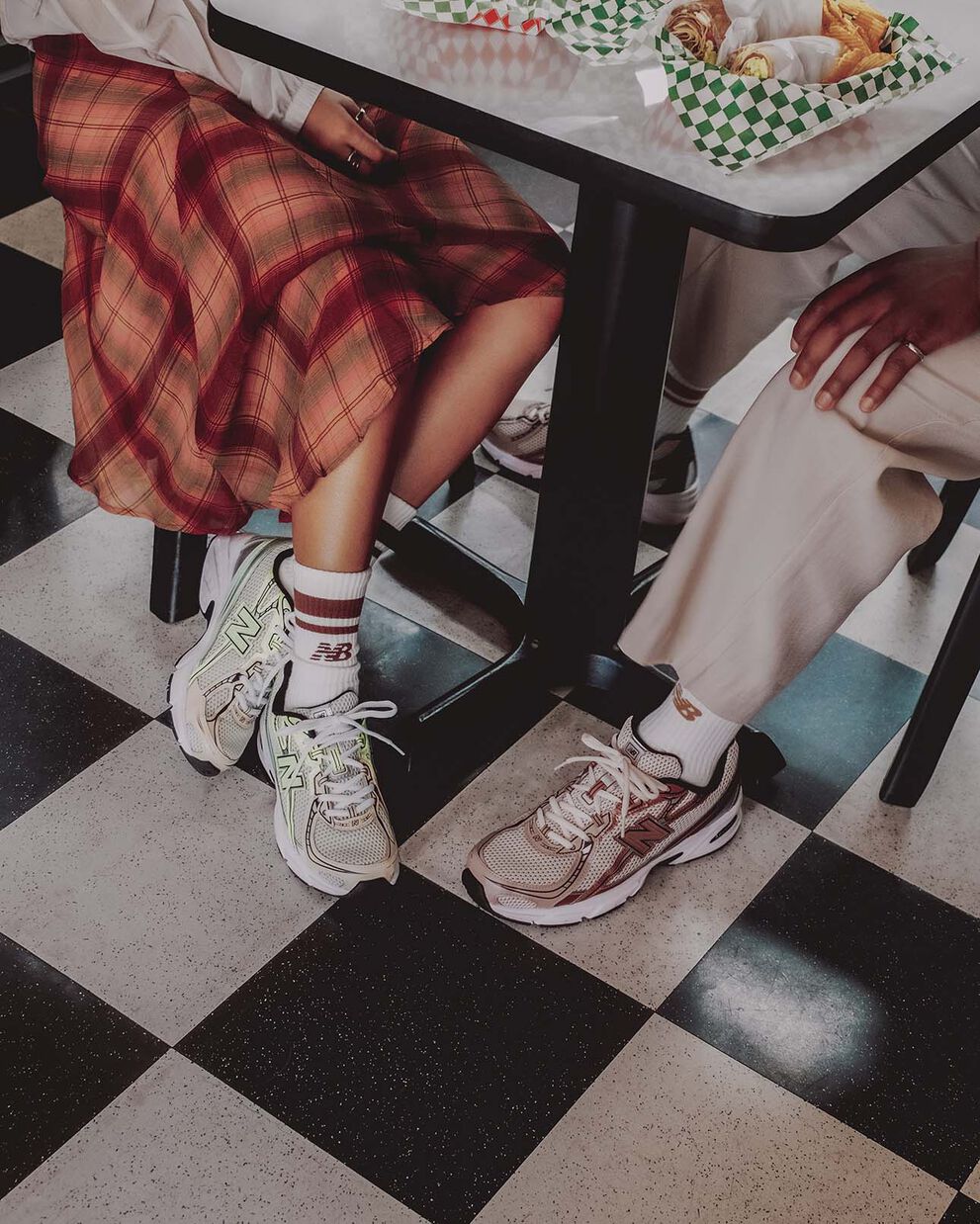 Knee-down image of a male and female model sitting at a table wearing the New Balance 740 sneakers in the brown metallic colorway and the mint flash green colorway.