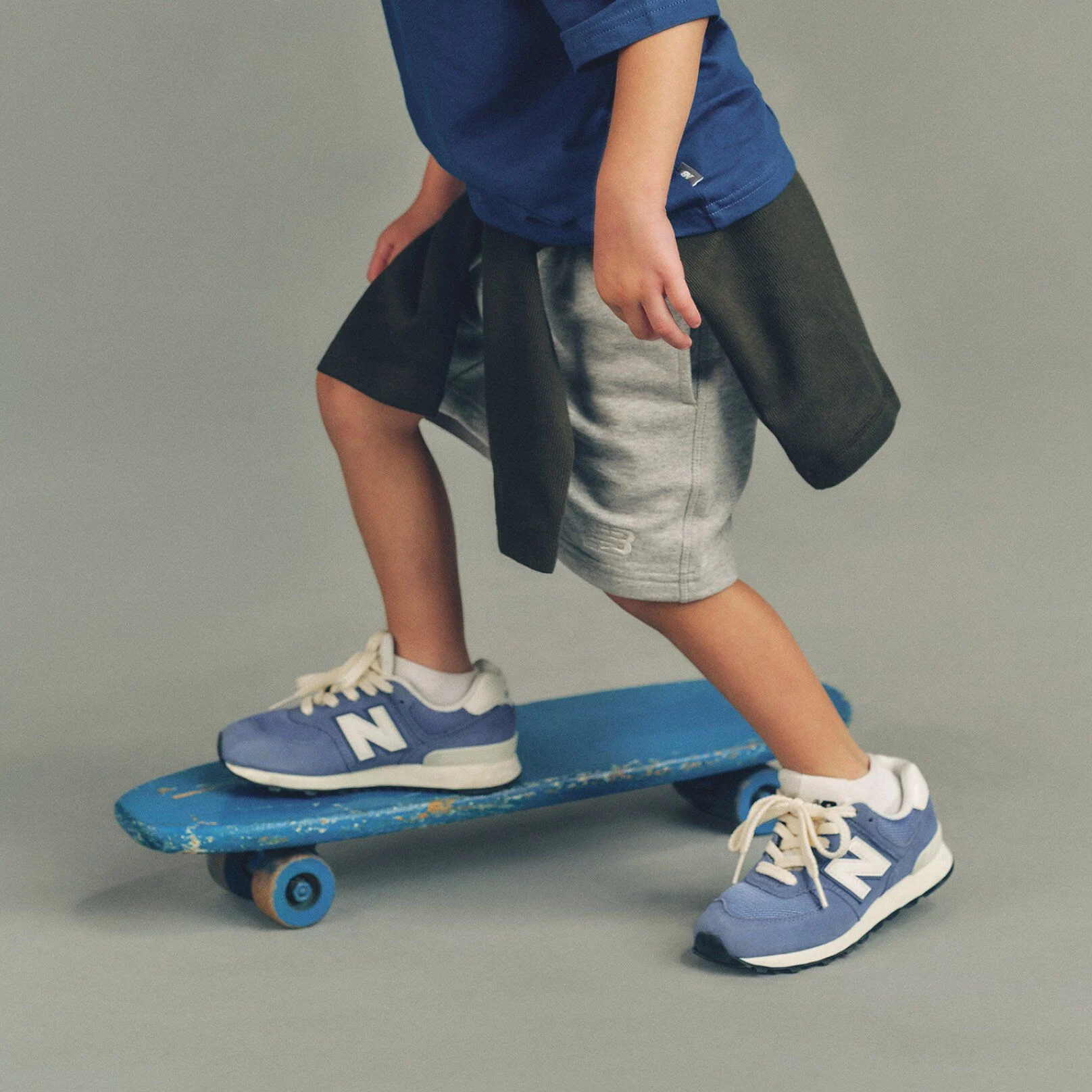 Studio image showing a child&rsquo;s lower body standing with one foot on a worn blue skateboard and the other foot on a smooth, light gray floor.