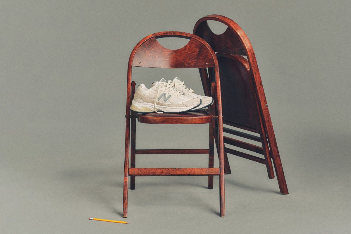 Studio image of a pair of cream-colored New Balance sneakers placed on a dark wooden folding chair, with a second matching chair folded and leaned behind it.