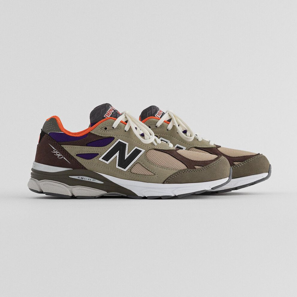 A pair of olive and brown 990v3 sneakers on a grey studio backdrop.