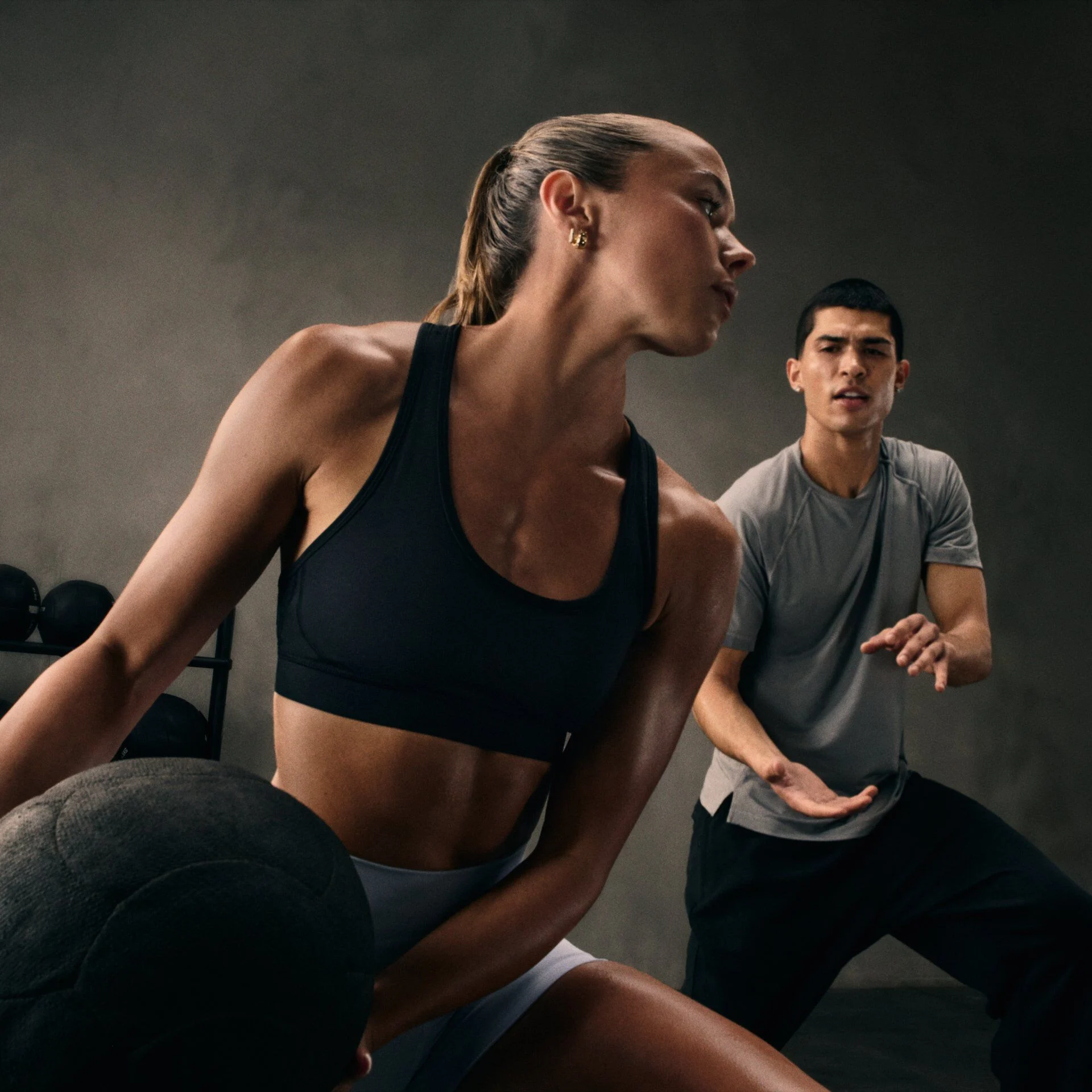 Two people exercising indoors, with one holding a medicine ball and another positioned behind in an athletic stance.