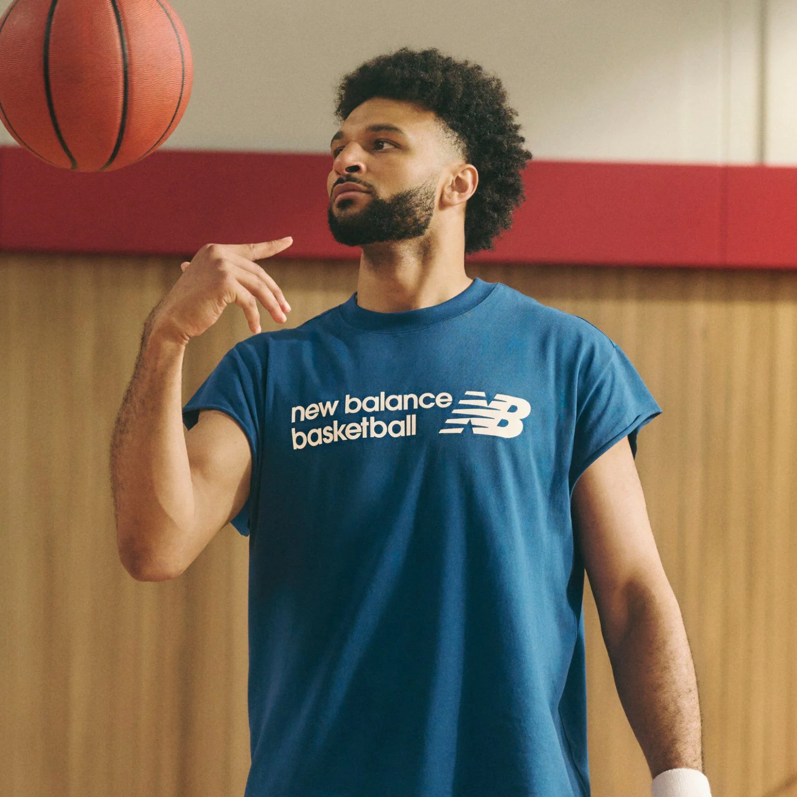 A person wearing a blue &ldquo;new balance basketball&rdquo; sleeveless T‑shirt casually spins a basketball on one hand while standing indoors against a wood-paneled and red-accented background.