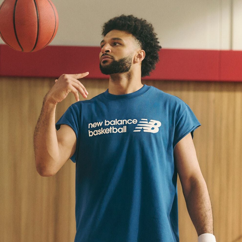 A person wearing a blue &ldquo;new balance basketball&rdquo; sleeveless T‑shirt casually spins a basketball on one hand while standing indoors against a wood-paneled and red-accented background.
