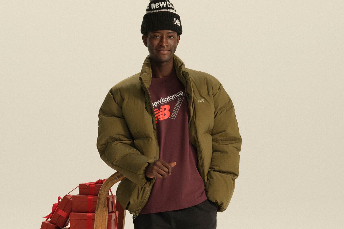 Person wearing an olive puffer jacket, burgundy New Balance shirt, and black knit beanie standing beside a wooden sled stacked with red gift boxes. 