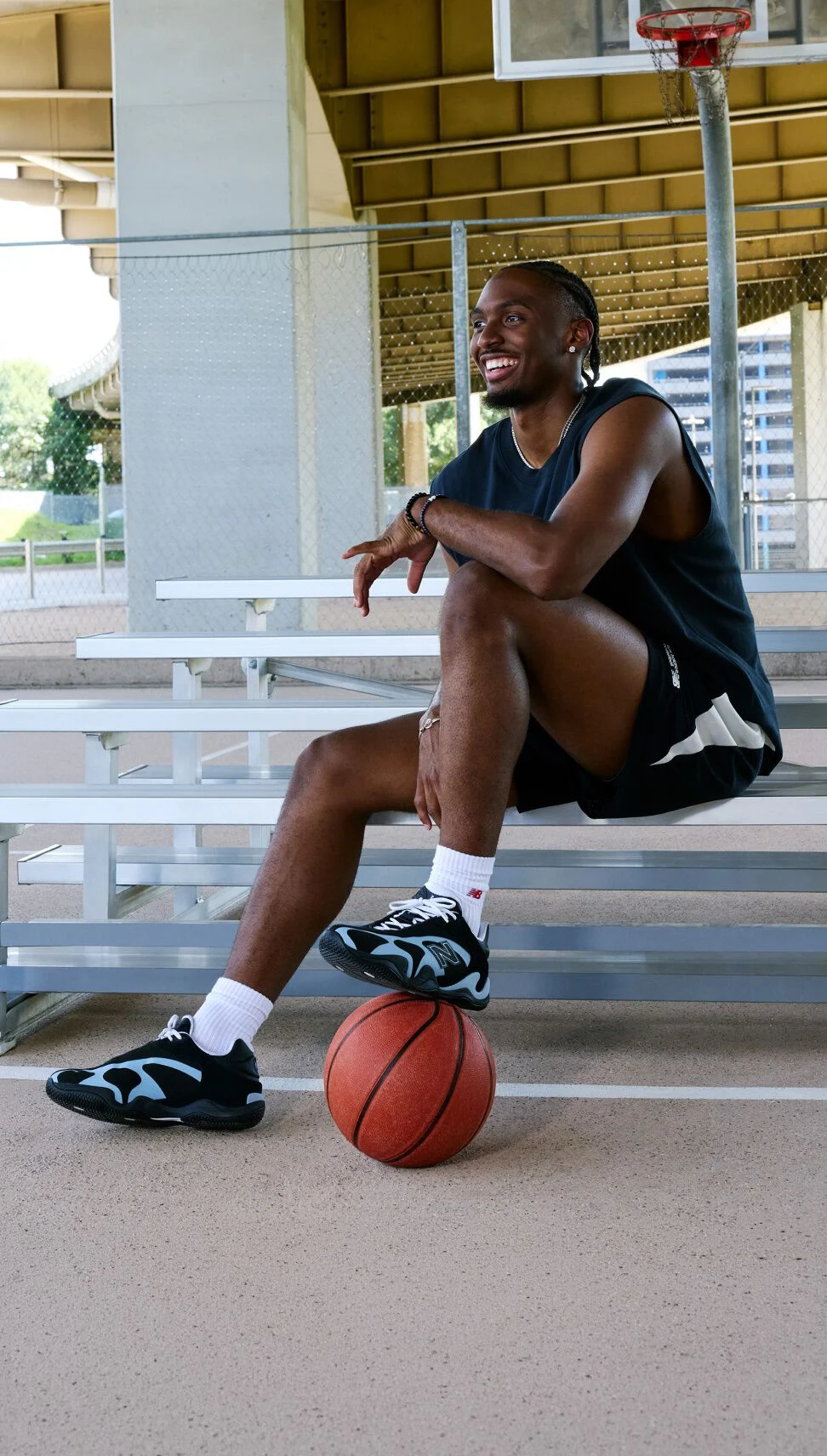 Person seated on outdoor bleachers beneath an overpass with one foot resting on a basketball, showcasing black-and-light-blue athletic sneakers.