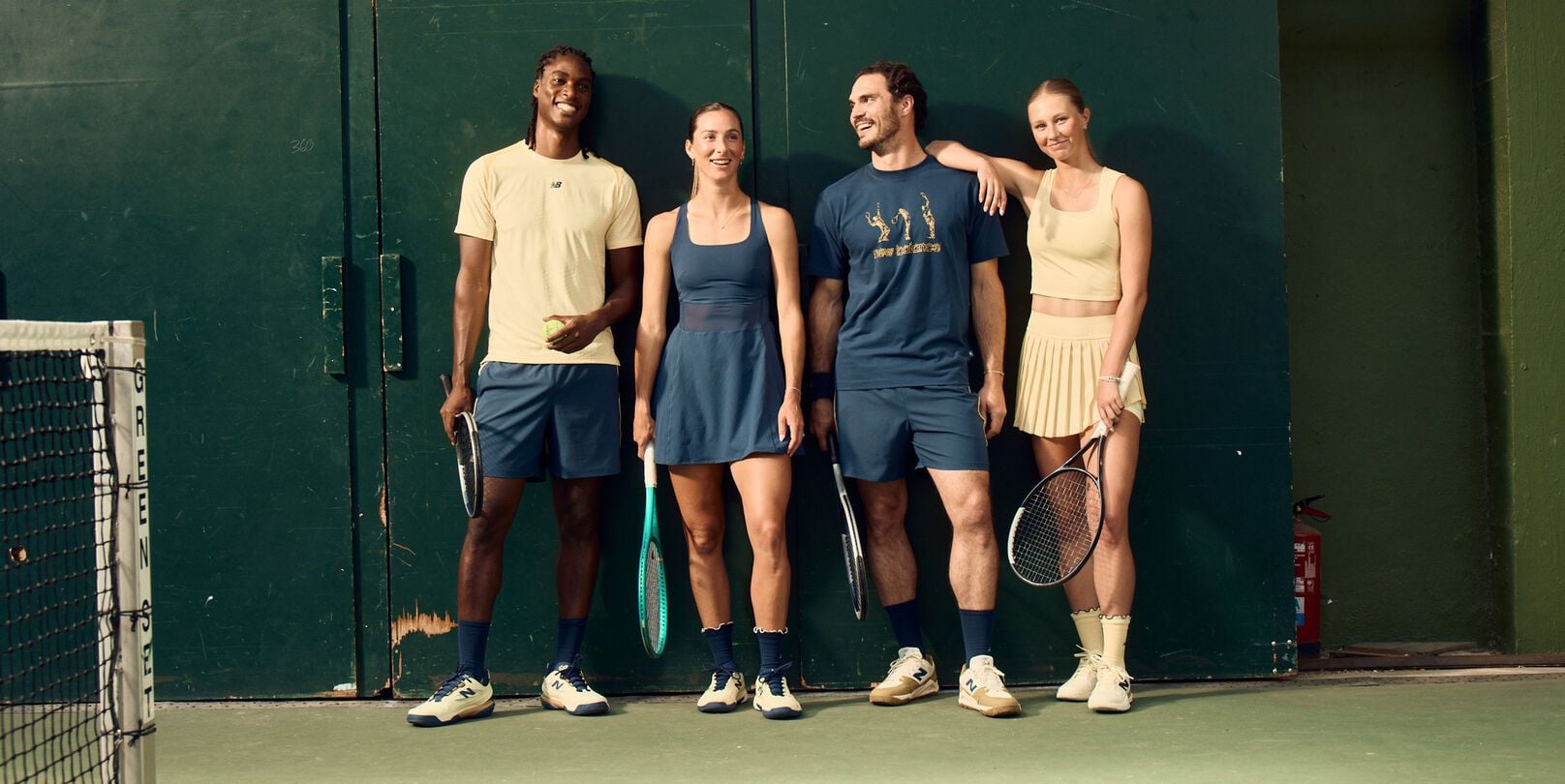 Four tennis players in coordinated athletic apparel stand against a dark green court wall holding rackets and a tennis ball.
