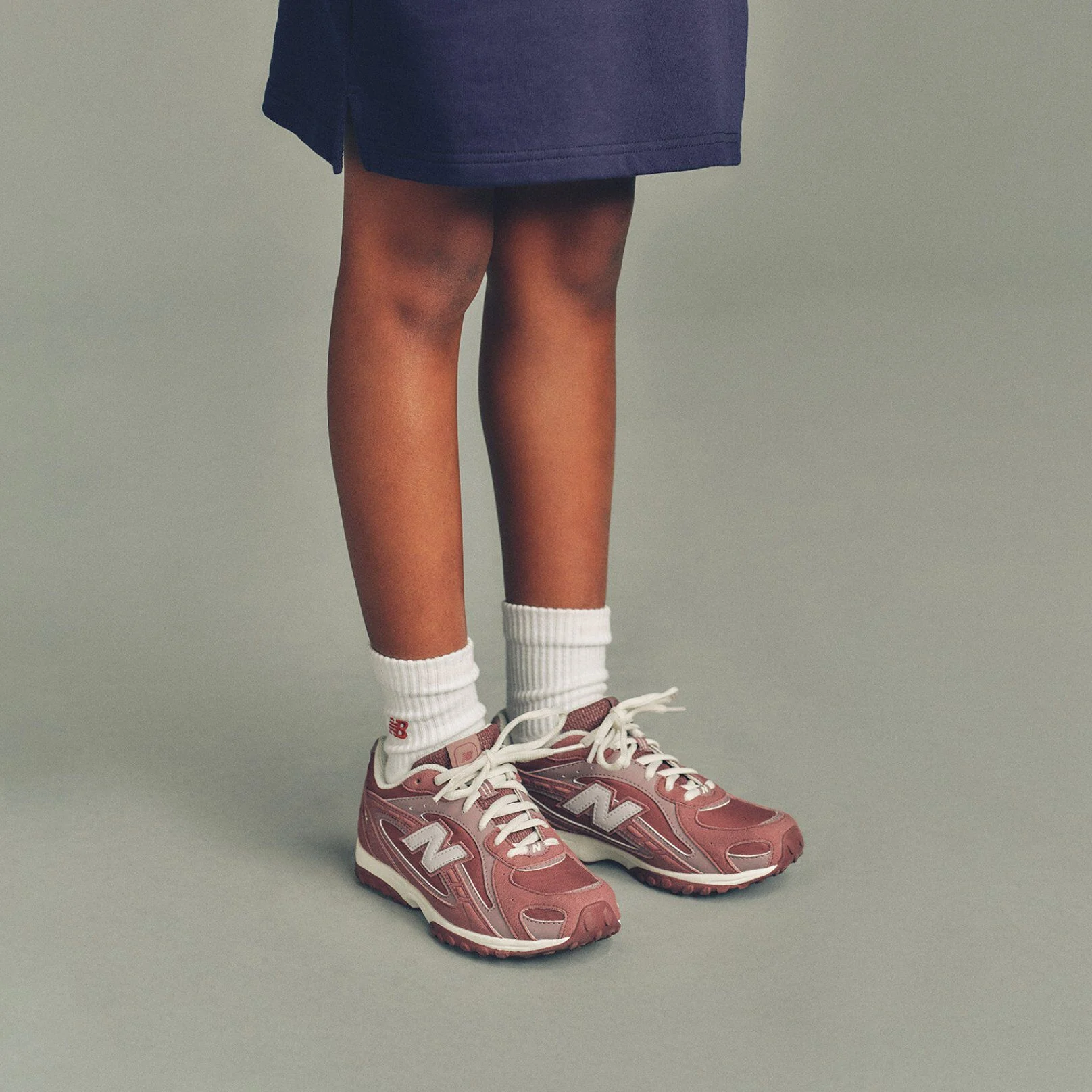 The child is wearing a navy top that extends to mid-thigh and white ribbed crew socks featuring a small red &ldquo;NB&rdquo; logo near the cuff