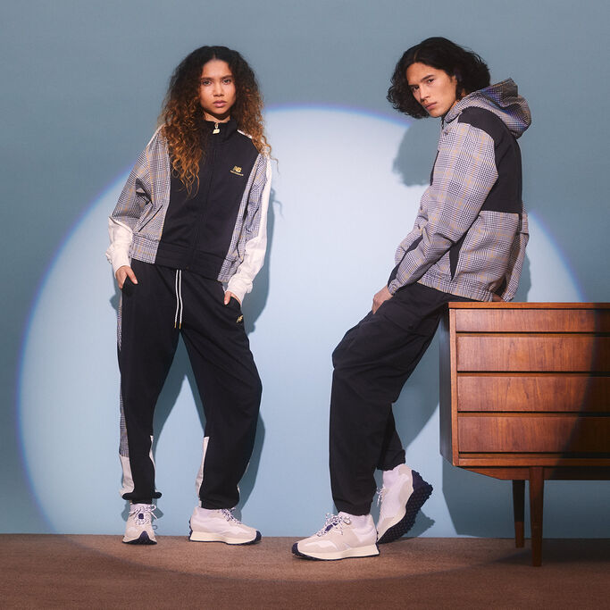 Man and women posed in NB Athletics gear and 327 shoes spotlighted in studio with vintage furniture and carpeting.