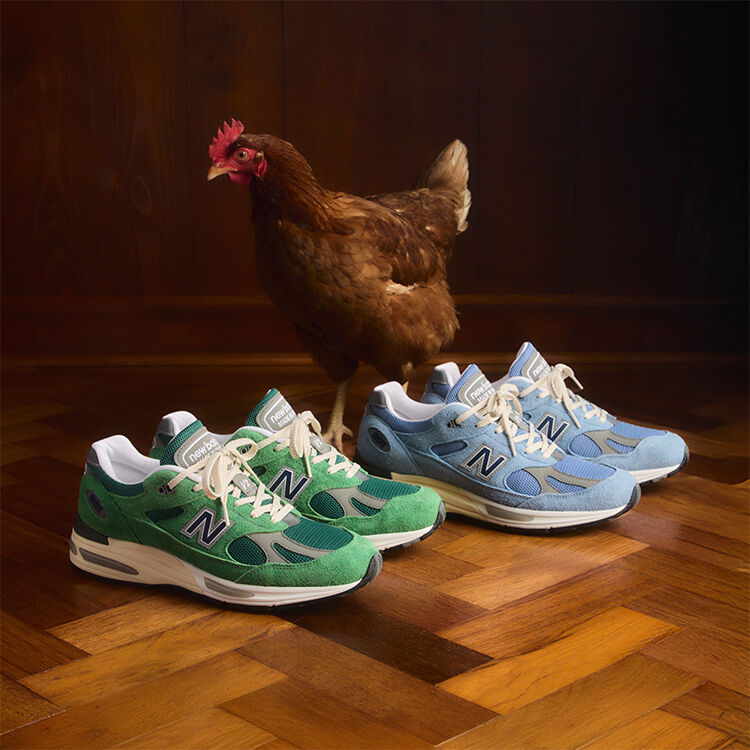 Two pairs of New Balance Made in UK sneakers sitting on a rustic herringbone wooden floor with a chicken standing behind them.  