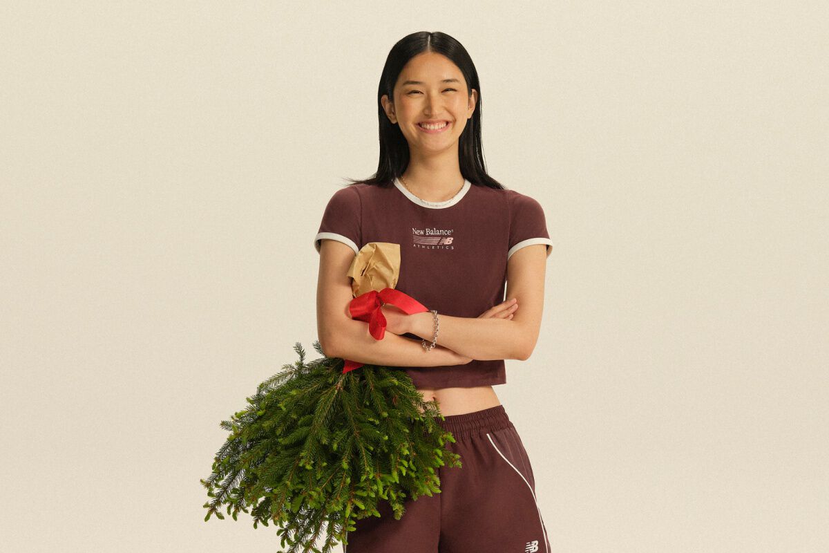 Person wearing a burgundy New Balance athletic set holding a wrapped gift and evergreen branches against a neutral studio background. 