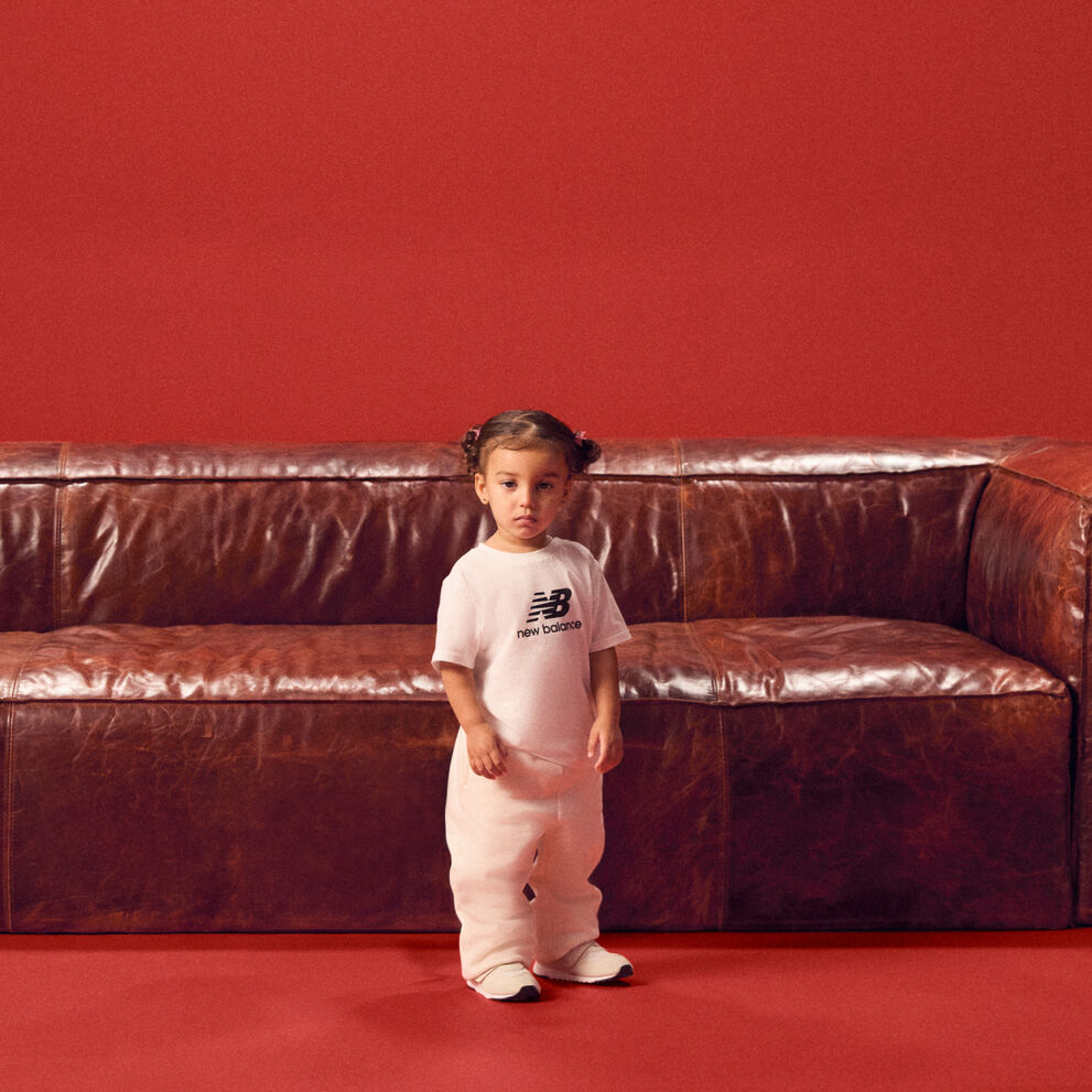 Baby standing in front of a red couch wearing a white tshirt.