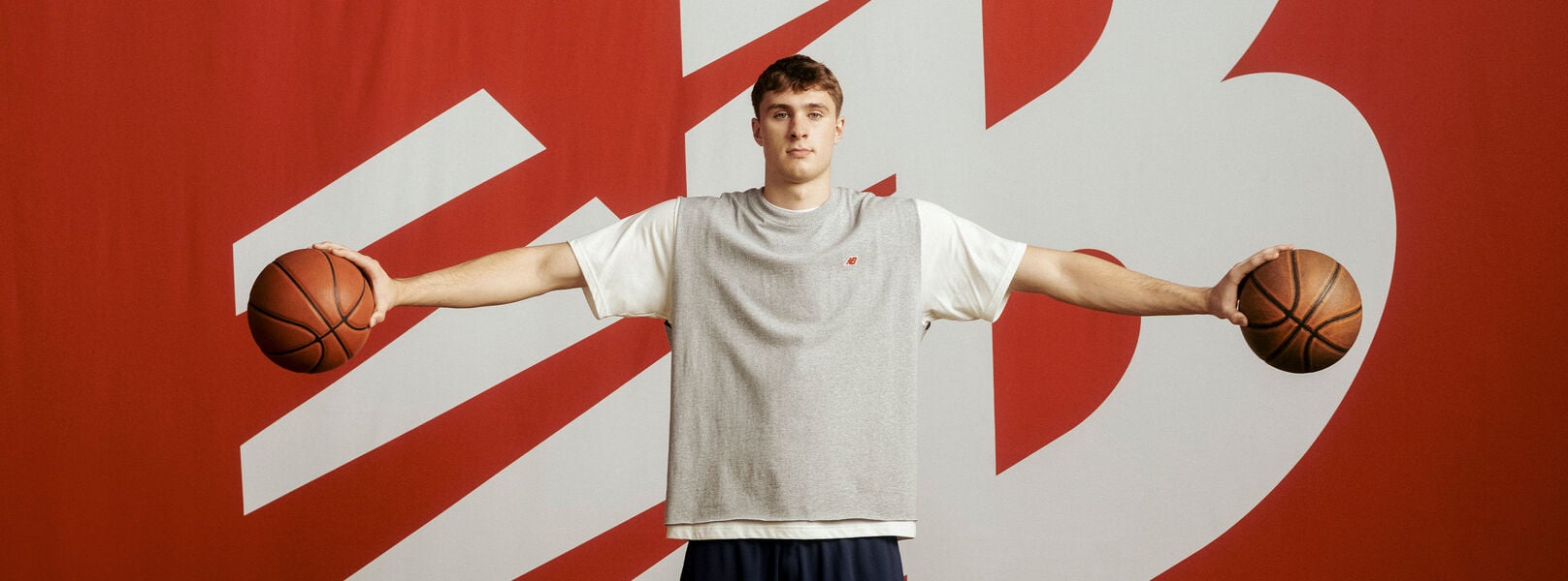 Cooper Flagg de pie frente a un gran fondo rojo con un logotipo blanco de New Balance, vistiendo una camiseta gris sin mangas y pantalones cortos azul marino, sosteniendo una pelota de baloncesto en cada mano con los brazos extendidos horizontalmente.