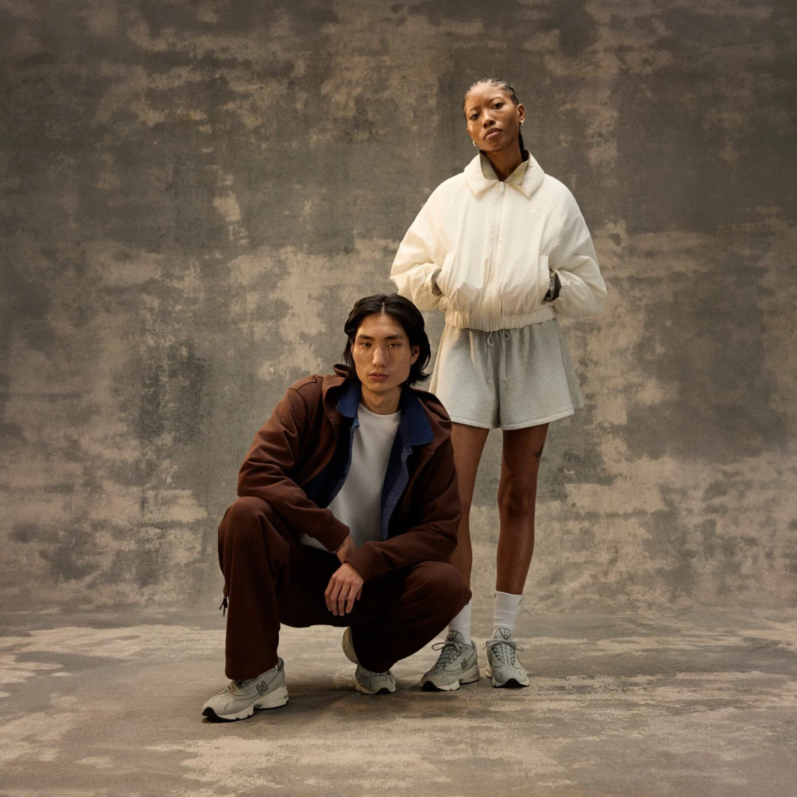 Two people posing indoors against a textured backdrop, wearing casual athletic outfits and sneakers.
