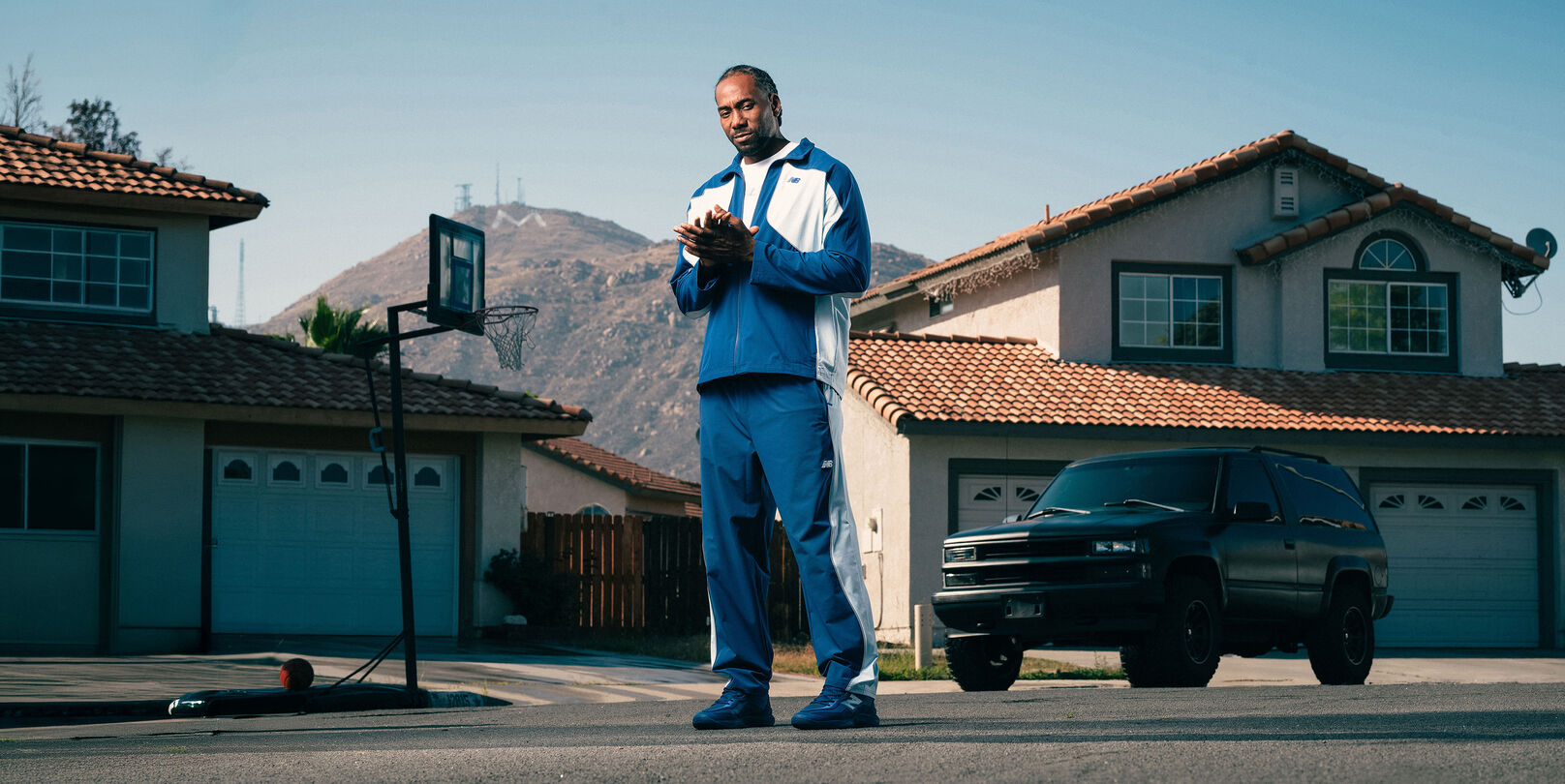 Person wearing a blue and white tracksuit and matching sneakers stands on a suburban street with mountains and houses in the background.