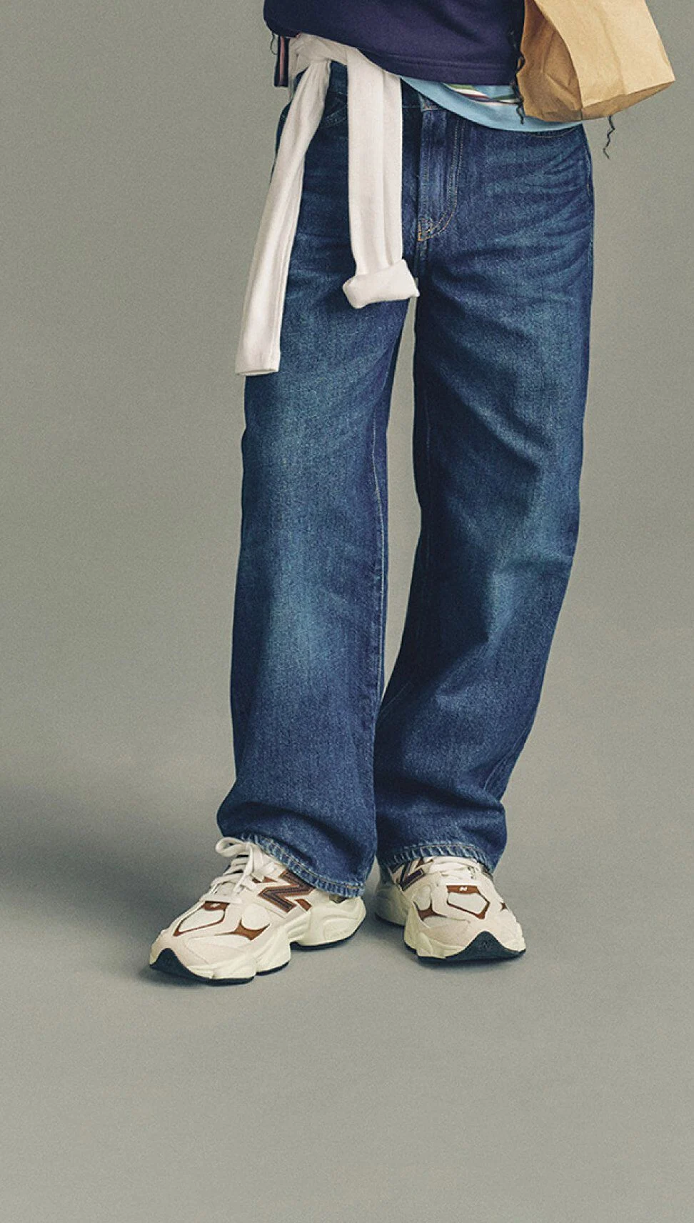 Close-up of a person wearing loose blue jeans and white New Balance sneakers with brown accents, standing against a plain background while holding a paper bag.