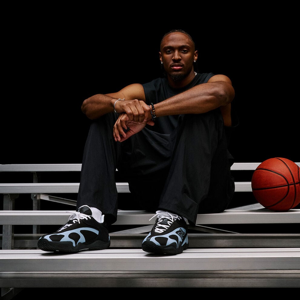 Person seated on metal bleachers beside a basketball, highlighting black-and-light-blue athletic sneakers in a dark studio setting.