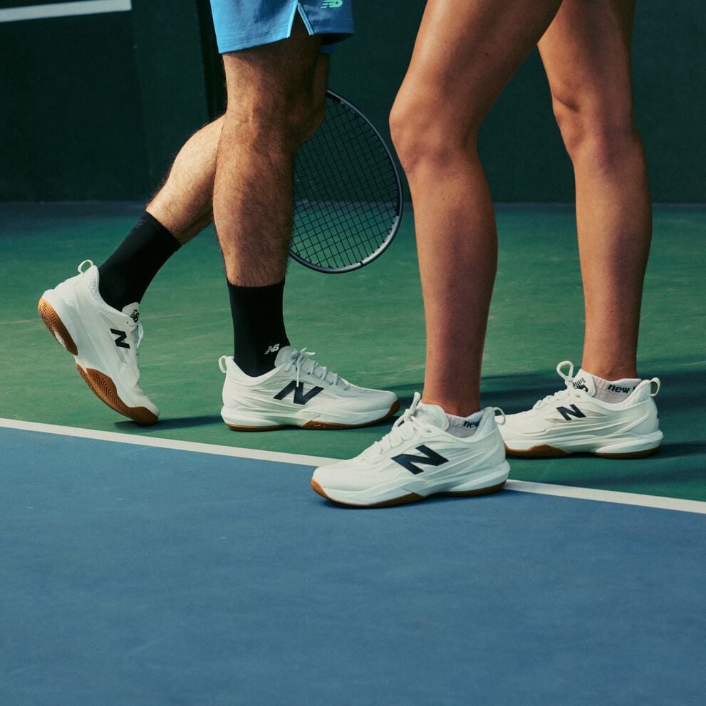 Primer plano de dos tenistas con zapatillas blancas de pista de pie sobre una pista cubierta con una raqueta visible al fondo.