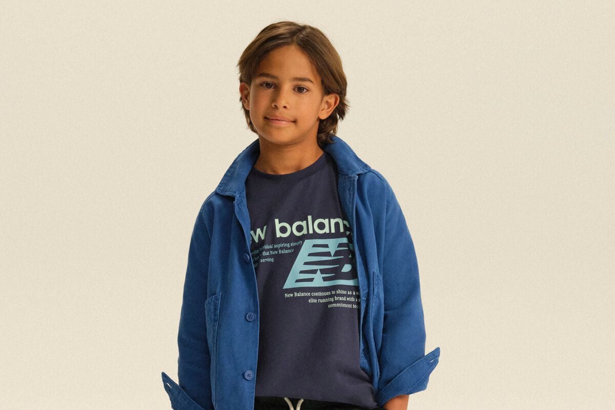 Child wearing a blue overshirt layered over a graphic New Balance T-shirt and dark joggers in a clean studio setting.