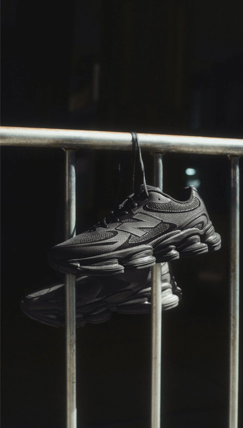 Black New Balance sneakers hanging by laces over a metal railing in dramatic lighting.