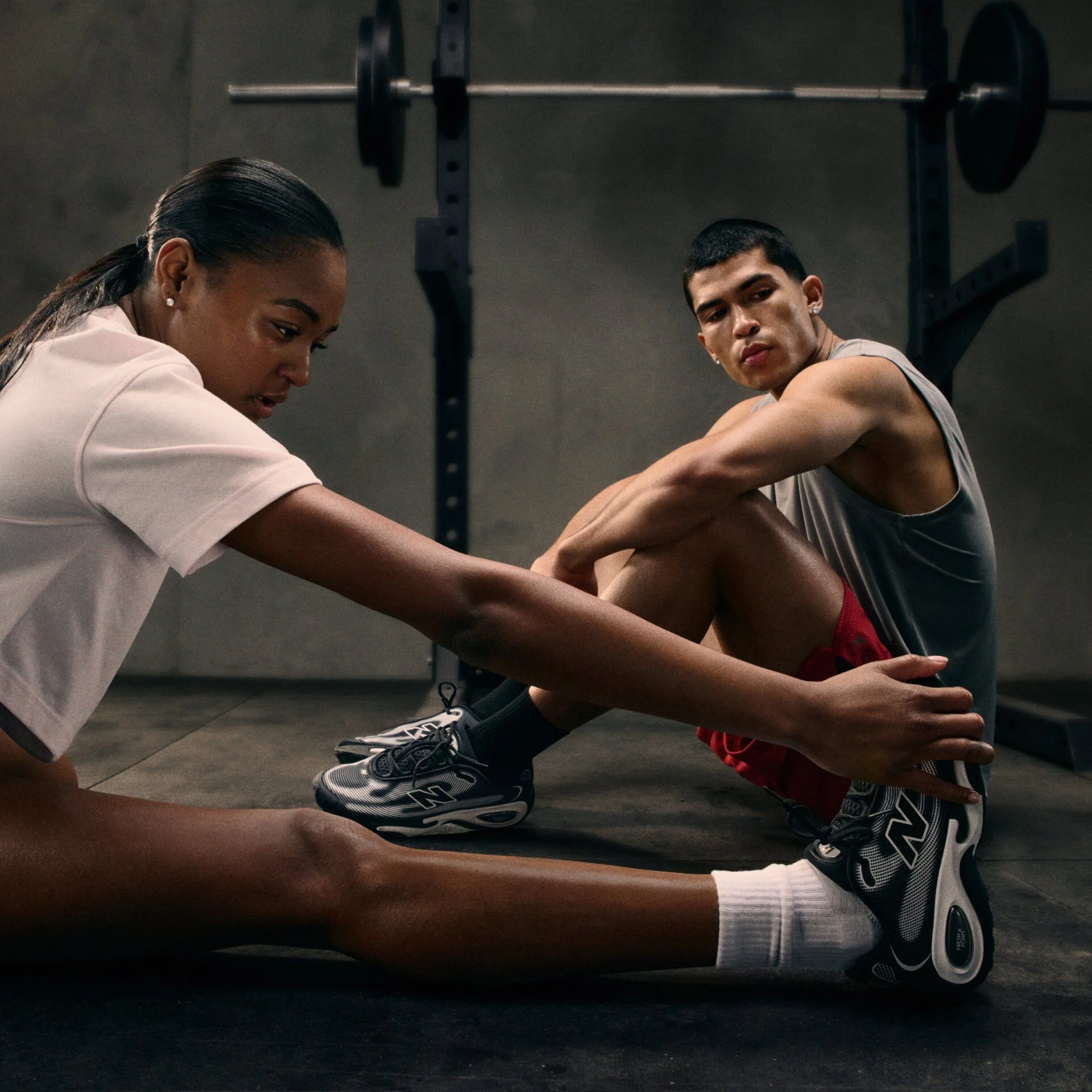 Two people seated on a gym floor stretching their legs, wearing athletic clothing and sneakers, with a barbell and squat rack visible in the background under low, moody lighting.