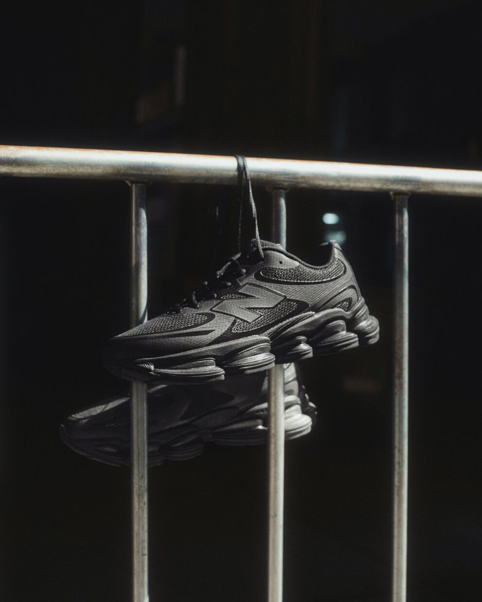 Black New Balance sneakers hanging by laces over a metal railing in dramatic lighting.