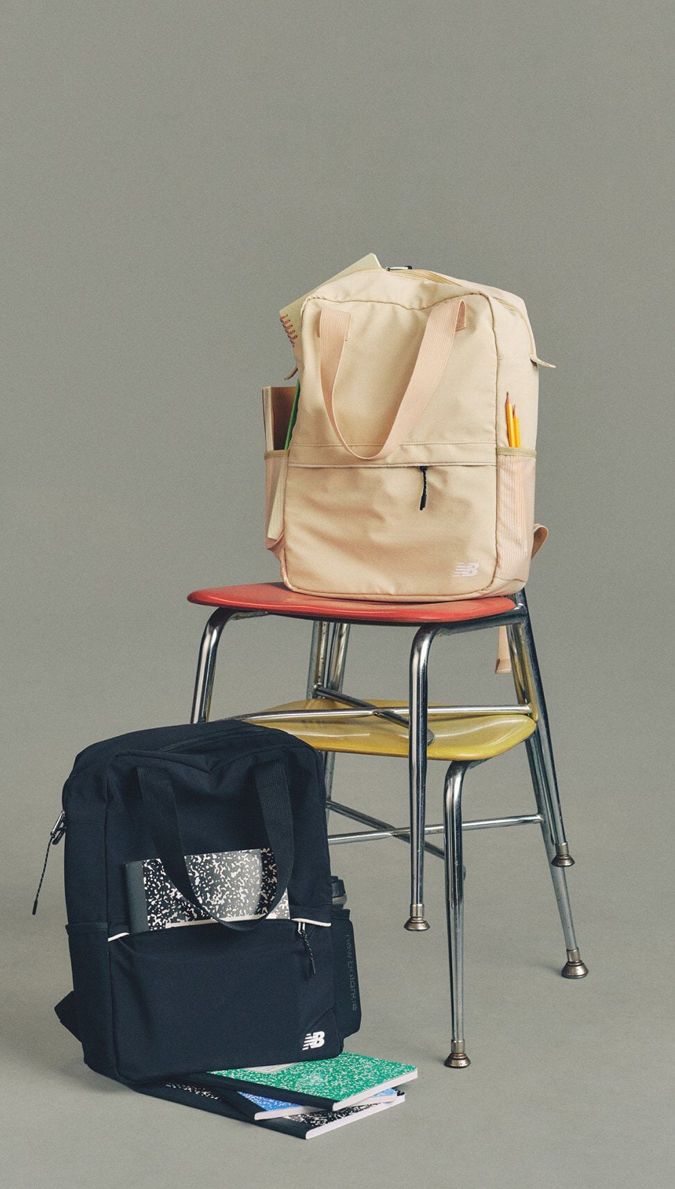 Imagen de estudio de dos mochilas New Balance &mdash;una beige sobre sillas apiladas y otra negra en el suelo&mdash; rodeadas de cuadernos y material escolar sobre un fondo gris neutro.