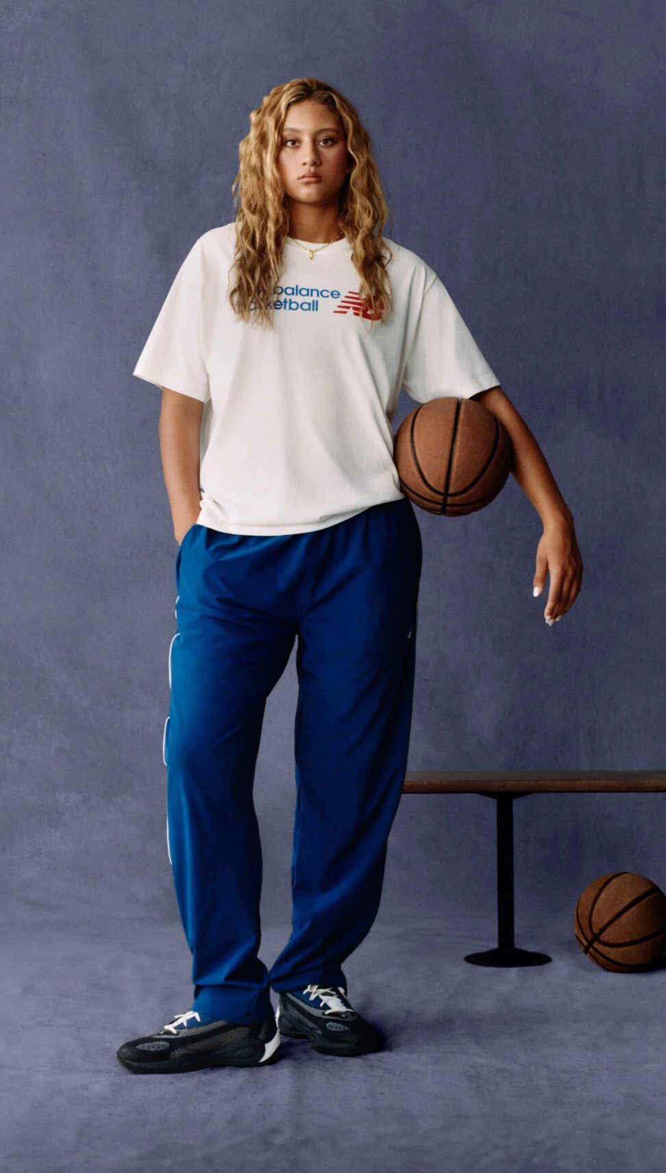 A person stands in a relaxed pose against a textured dark backdrop, wearing a white New Balance Basketball T‑shirt and blue athletic pants.