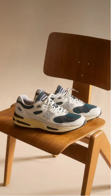 A pair of New Balance Made in UK sneakers sitting on a wooden chair in a beige studio environment. 
